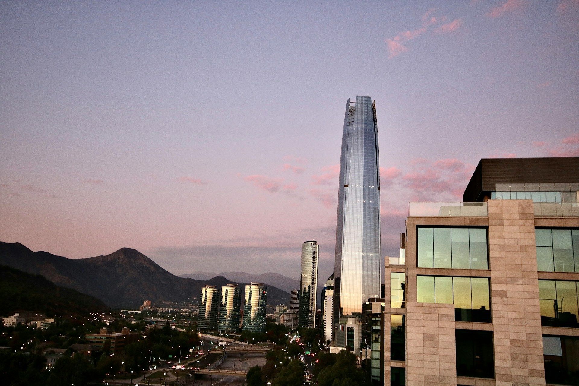 Uno skyline cittadino moderno con diversi grattacieli che si stagliano su una catena montuosa sotto un cielo rosa e viola al tramonto.