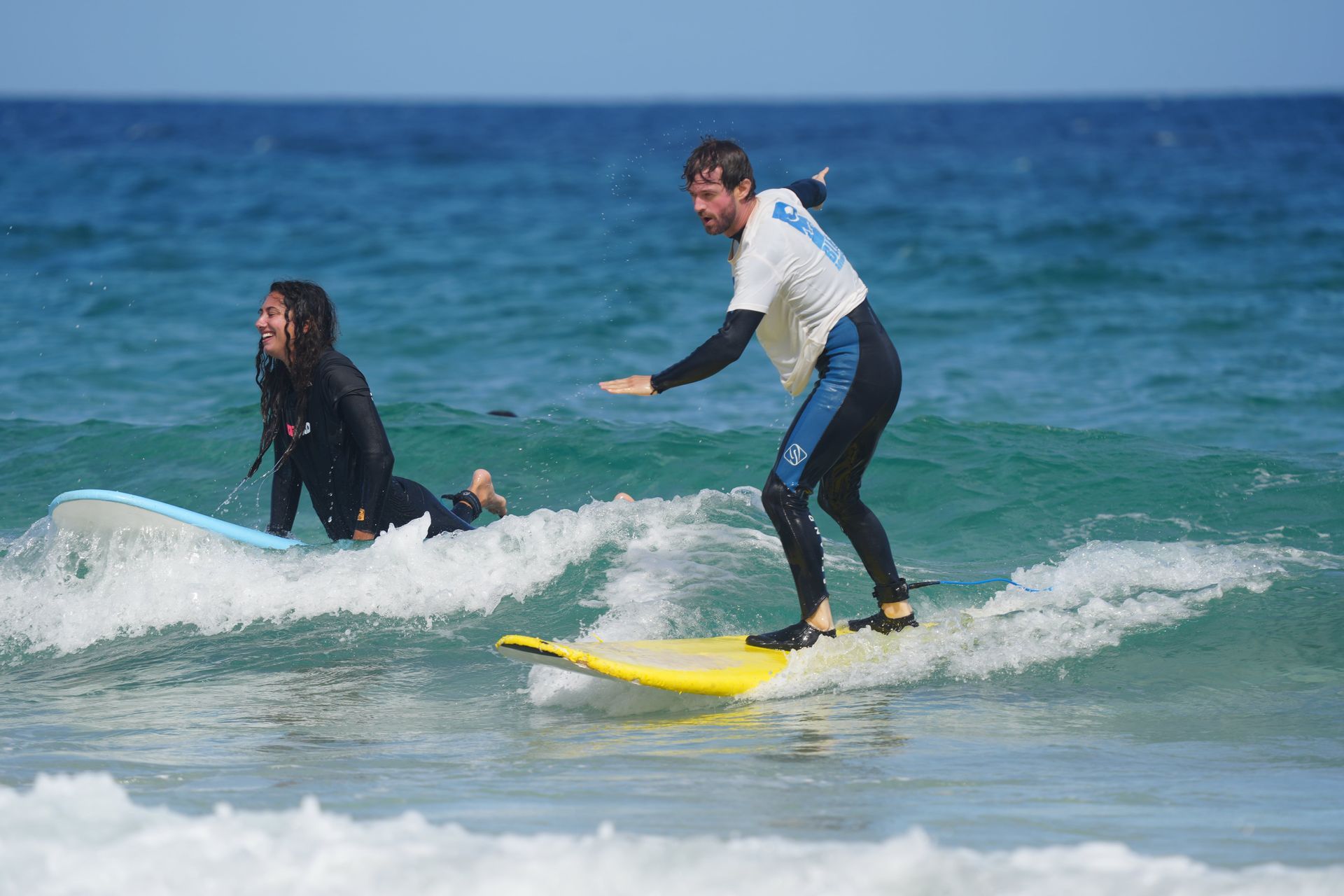 Un homme en voyage de groupe WeRoad fait de l'équilibre sur une planche de surf jaune tandis qu'une femme sur une autre planche sourit dans l'océan.