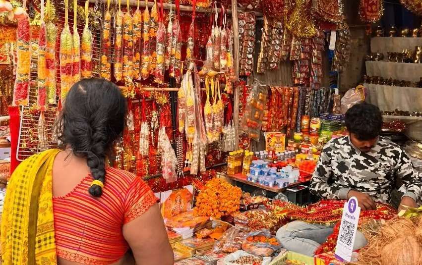 Una donna in sari osserva una bancarella in un mercato all'aperto, piena di ghirlande colorate e articoli religiosi venduti da un uomo.