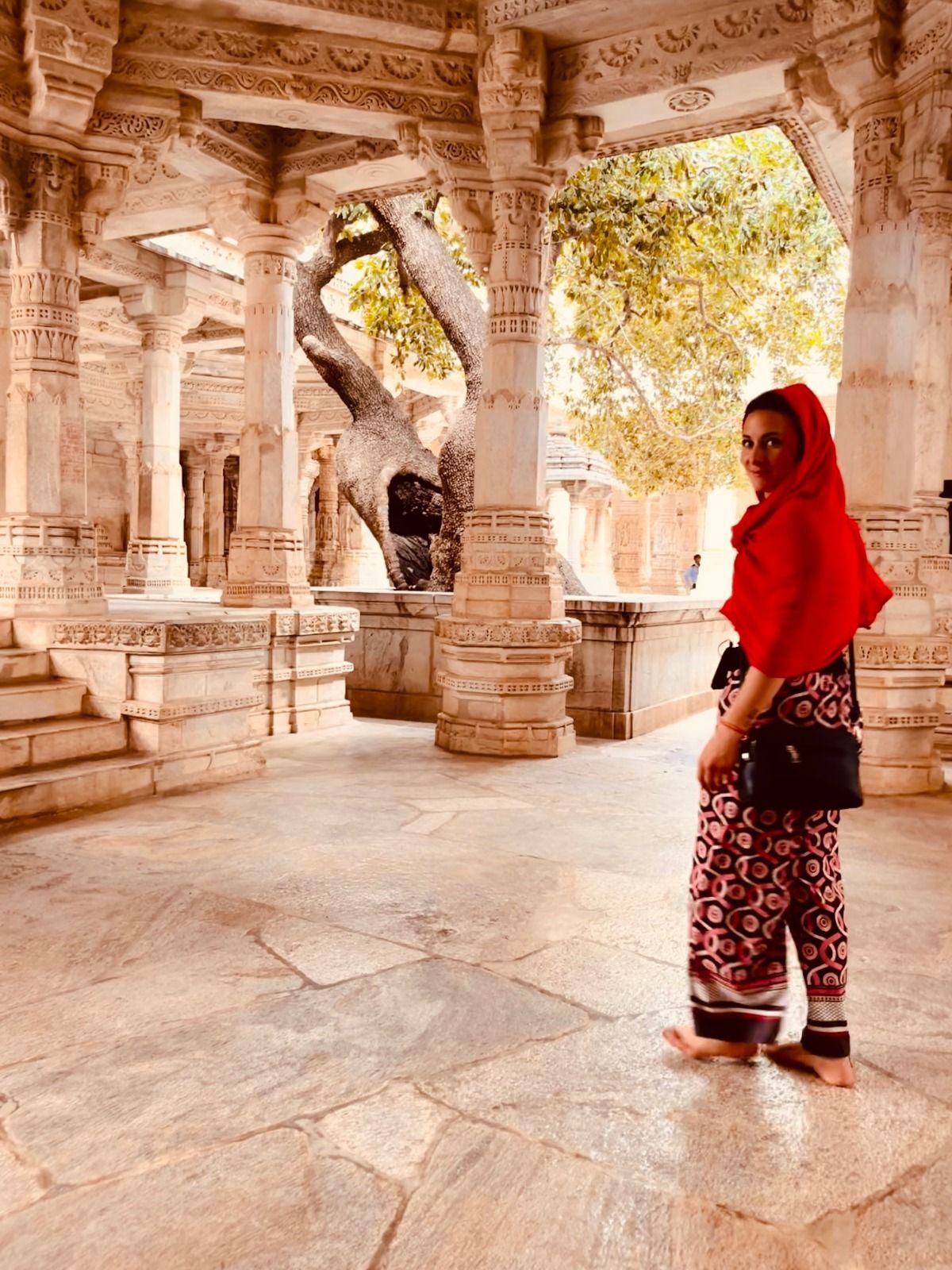 Una donna a piedi nudi con un foulard rosso si guarda indietro mentre si trova in un antico tempio con colonne di pietra intagliate e ornate.