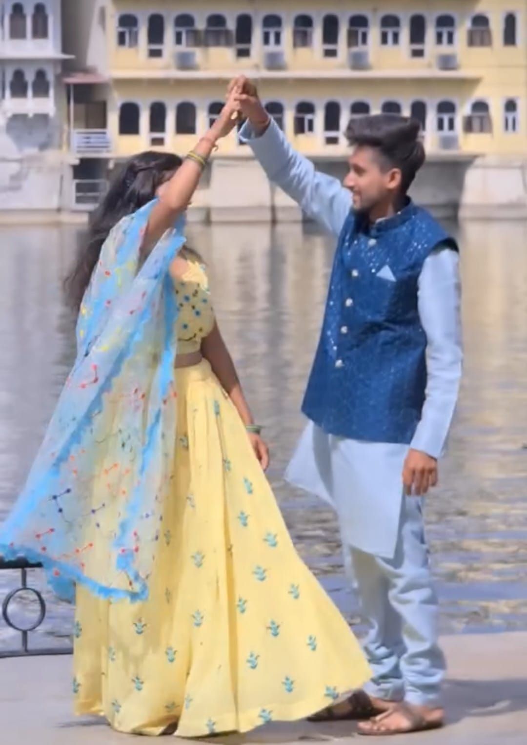 Un uomo e una donna in abiti tradizionali indiani ballano insieme, tenendosi per mano vicino a uno specchio d'acqua con un edificio sullo sfondo.
