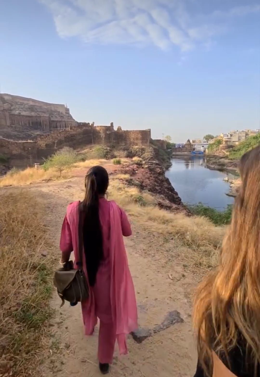 Due donne in un viaggio di gruppo WeRoad camminano su un sentiero sterrato lungo un fiume, con antiche fortificazioni in pietra sullo sfondo.