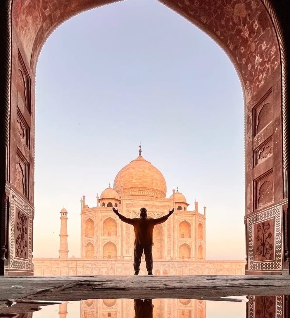 Una persona in silhouette a braccia alzate si erge davanti al Taj Mahal, visto attraverso un arco ornato con un riflesso in primo piano.