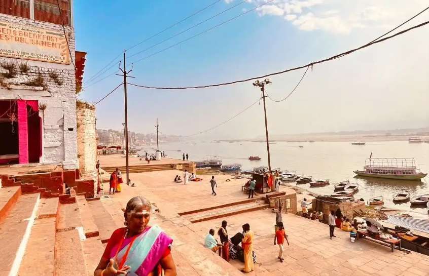 Una donna anziana in un sari colorato sale dei gradini di pietra che si affacciano su un ampio fiume con barche e persone sulla riva.