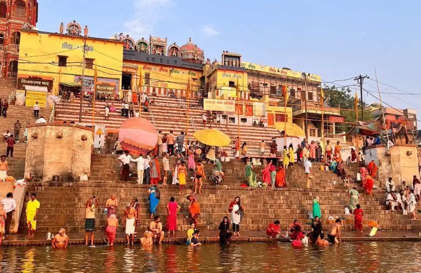 Una folla di persone vestite con abiti colorati si raduna su gradini di pietra accanto a un fiume, con alcuni che fanno il bagno nell'acqua.