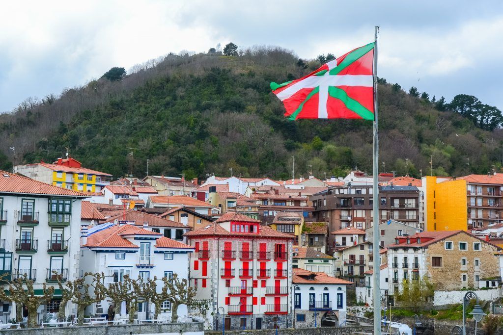 The Basque flag waves on a flagpole over colorful buildings at the base of a tree-covered hill under a cloudy sky.