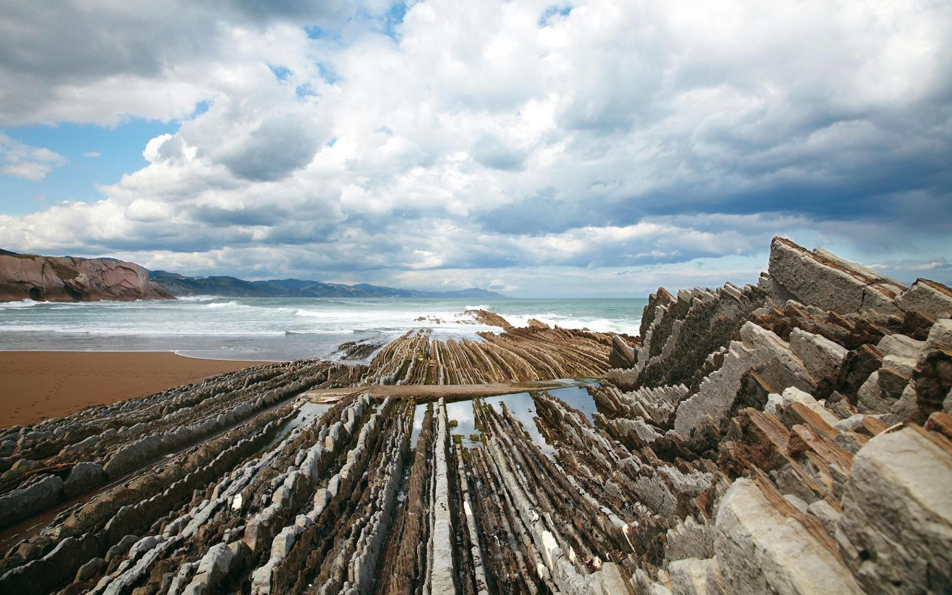 Layers of jagged rock formations extend from a sandy beach into the ocean under a cloudy sky.