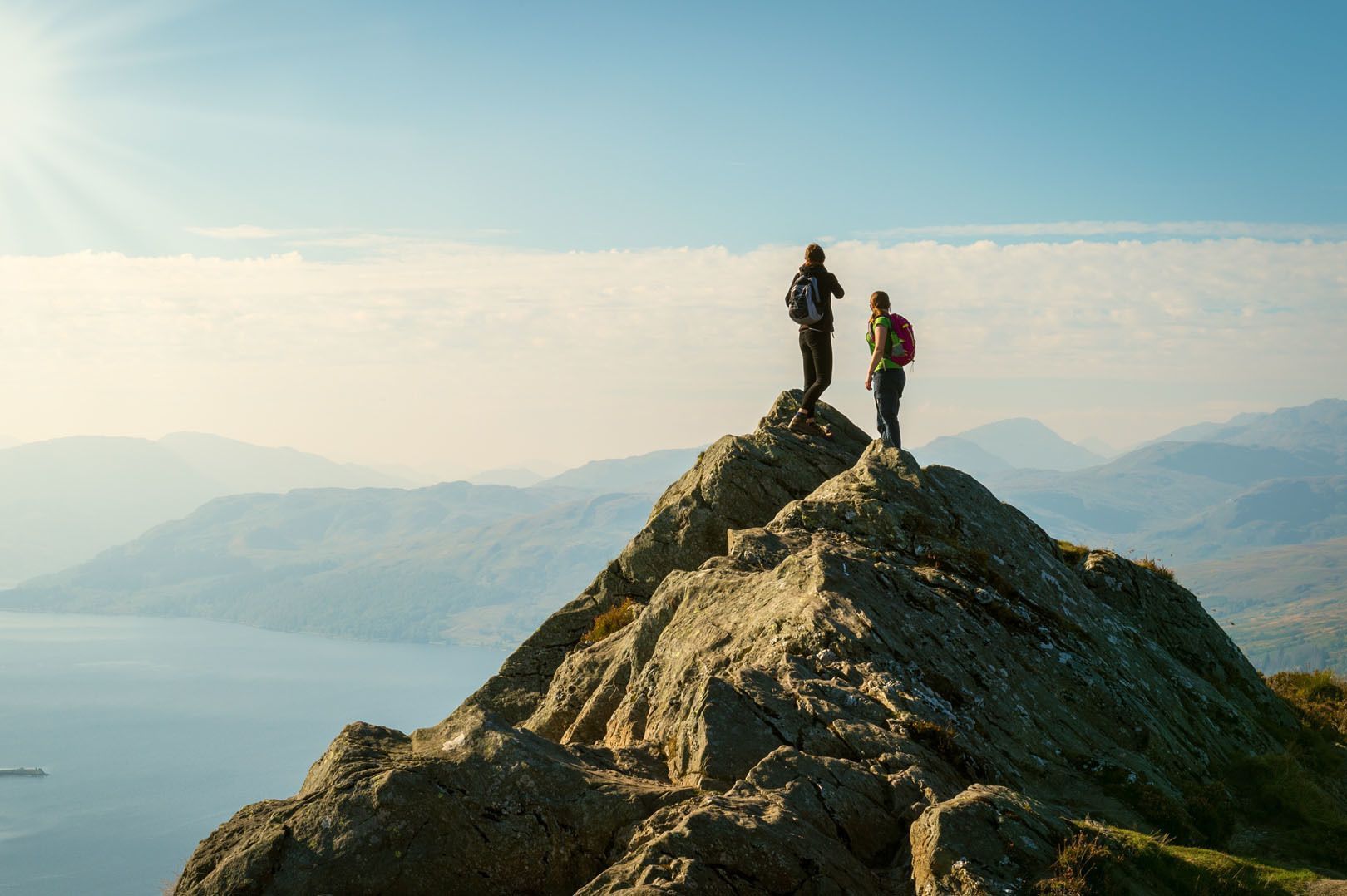Dos excursionistas de un viaje en grupo de WeRoad están de pie en la cima rocosa de una montaña, contemplando un vasto paisaje de montañas y un lago.