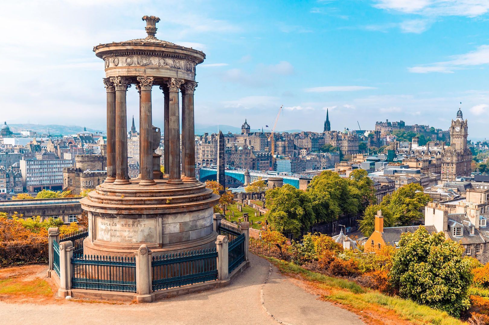 Un monumento clásico de piedra en una colina con vistas a un paisaje urbano histórico lleno de árboles de colores otoñales bajo un cielo azul.