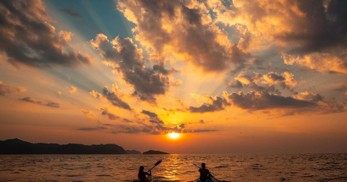 Due persone in kayak sul mare al tramonto, con i raggi del sole che filtrano tra nuvole arancioni e blu.