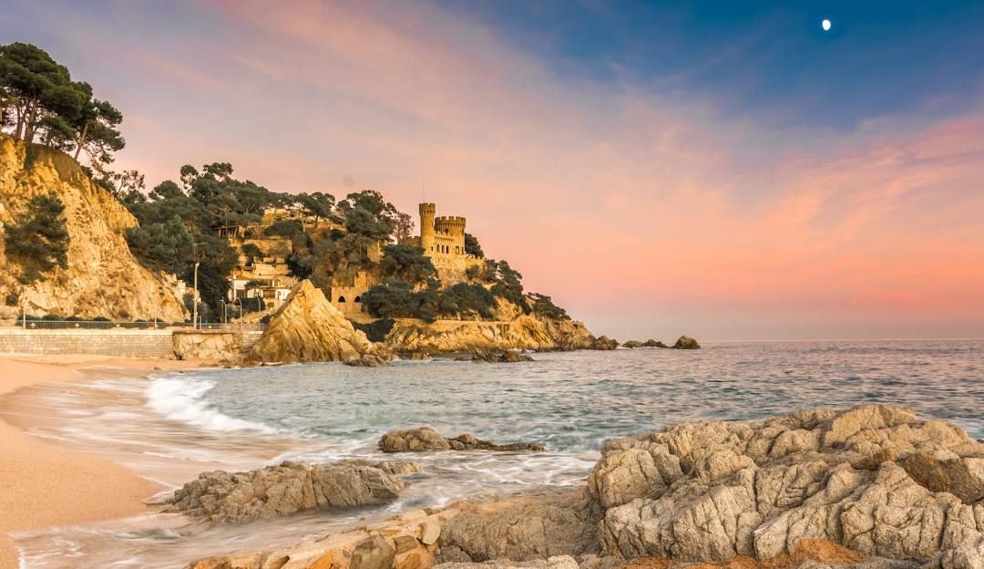 Un castello si erge su una scogliera rocciosa e alberata che si affaccia su una spiaggia sabbiosa e sull'oceano durante un tramonto colorato con la luna nel cielo.
