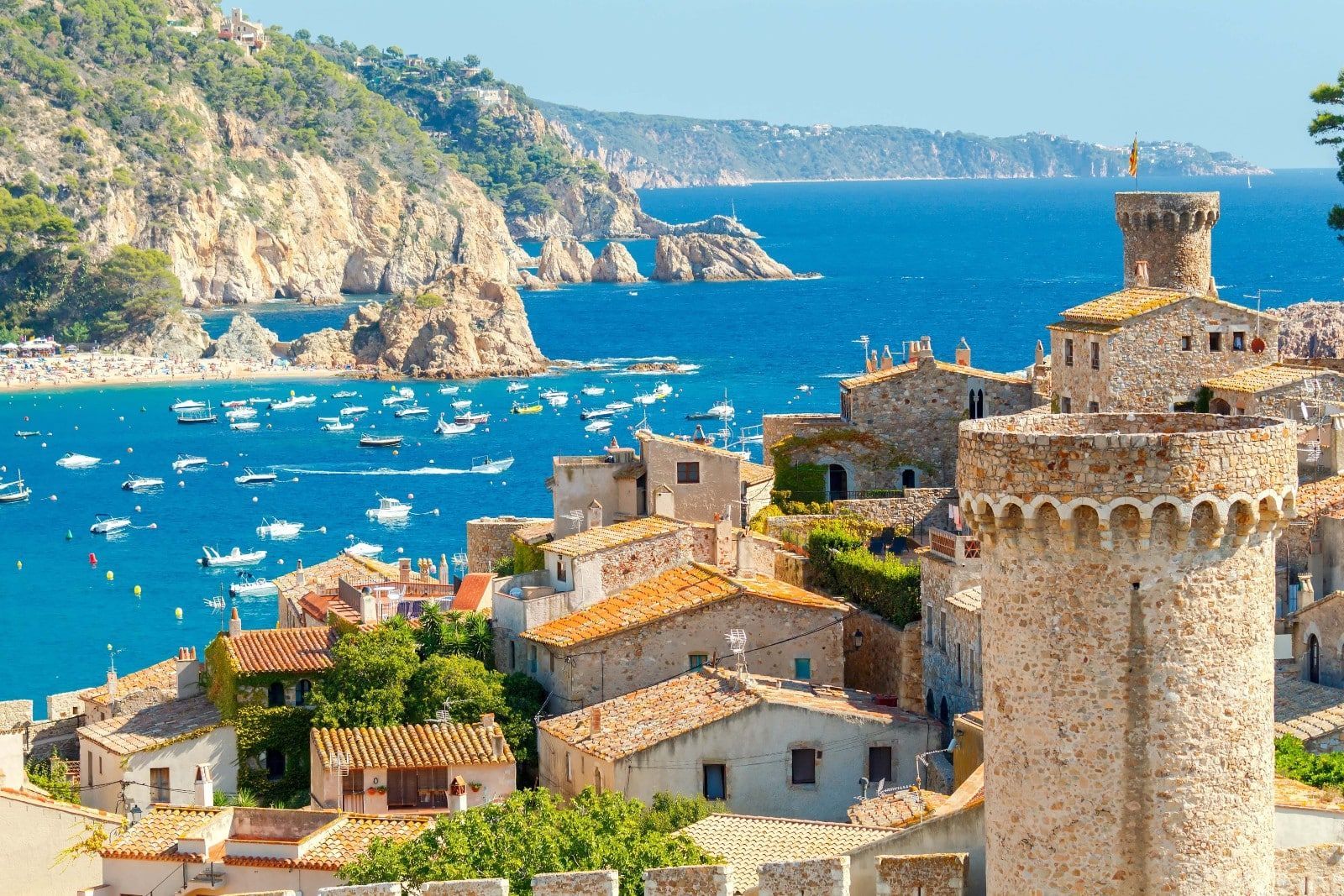 Una cittadina costiera, con edifici in pietra e una torre, si affaccia su una baia azzurra, piena di piccole imbarcazioni bianche e una spiaggia a ridosso della scogliera.