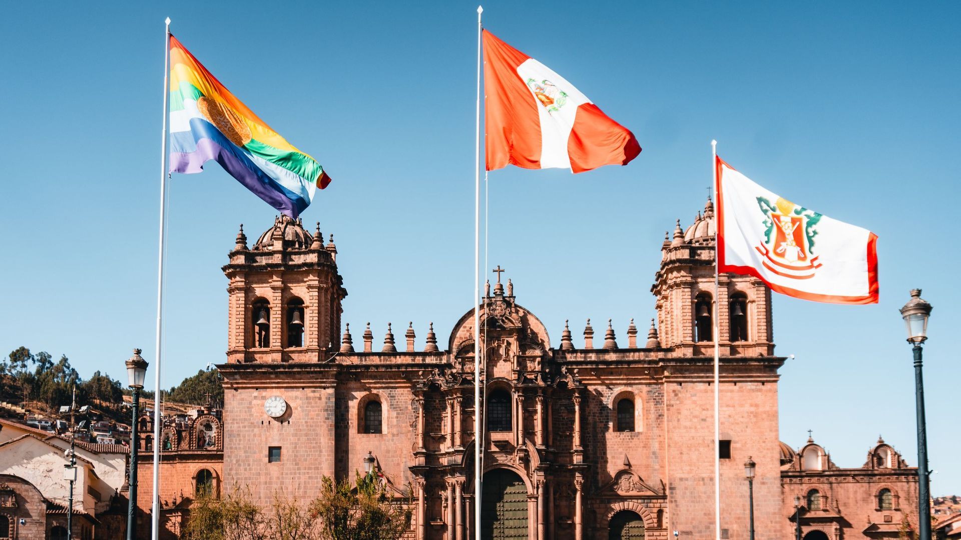 A rainbow flag, the flag of Peru, and the flag of Cusco waving on flagpoles in front of a large stone cathedral.