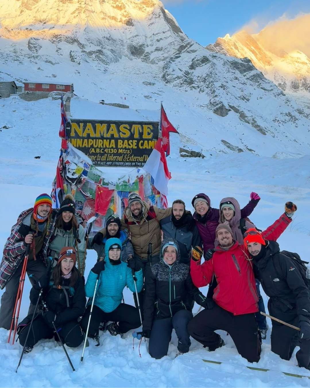 Un viaje en grupo de WeRoad posa para una foto en la nieve frente al cartel del Campo Base Annapurna, con un pico de montaña al fondo.