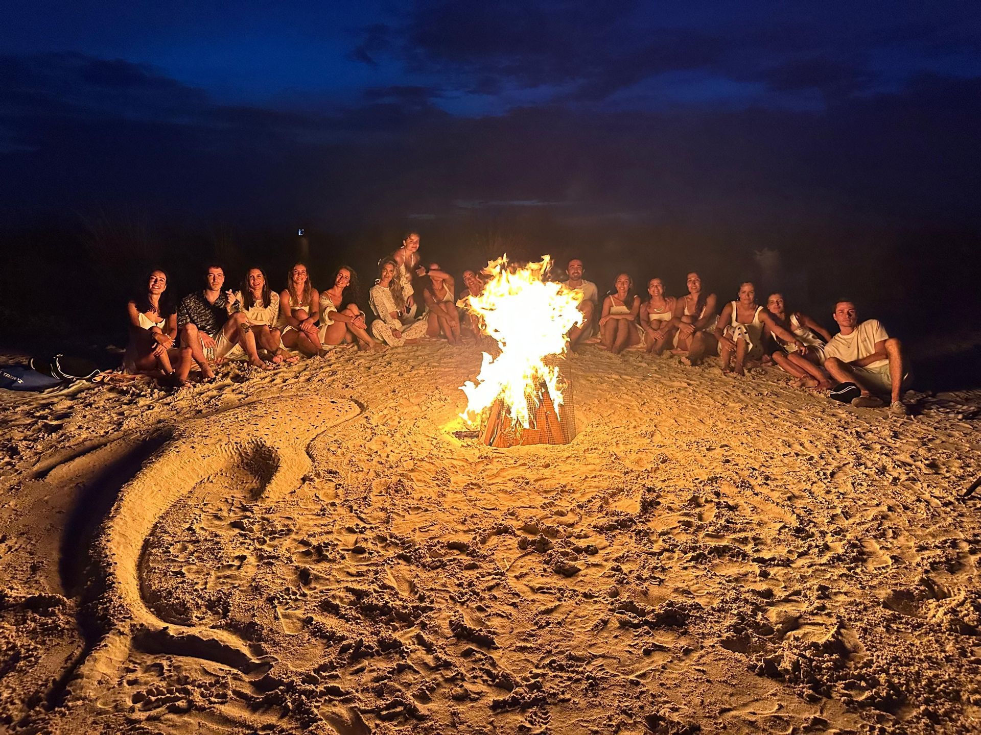 Un gruppo WeRoad riunito attorno a un falò su una spiaggia di notte, con un grande delfino disegnato sulla sabbia in primo piano.