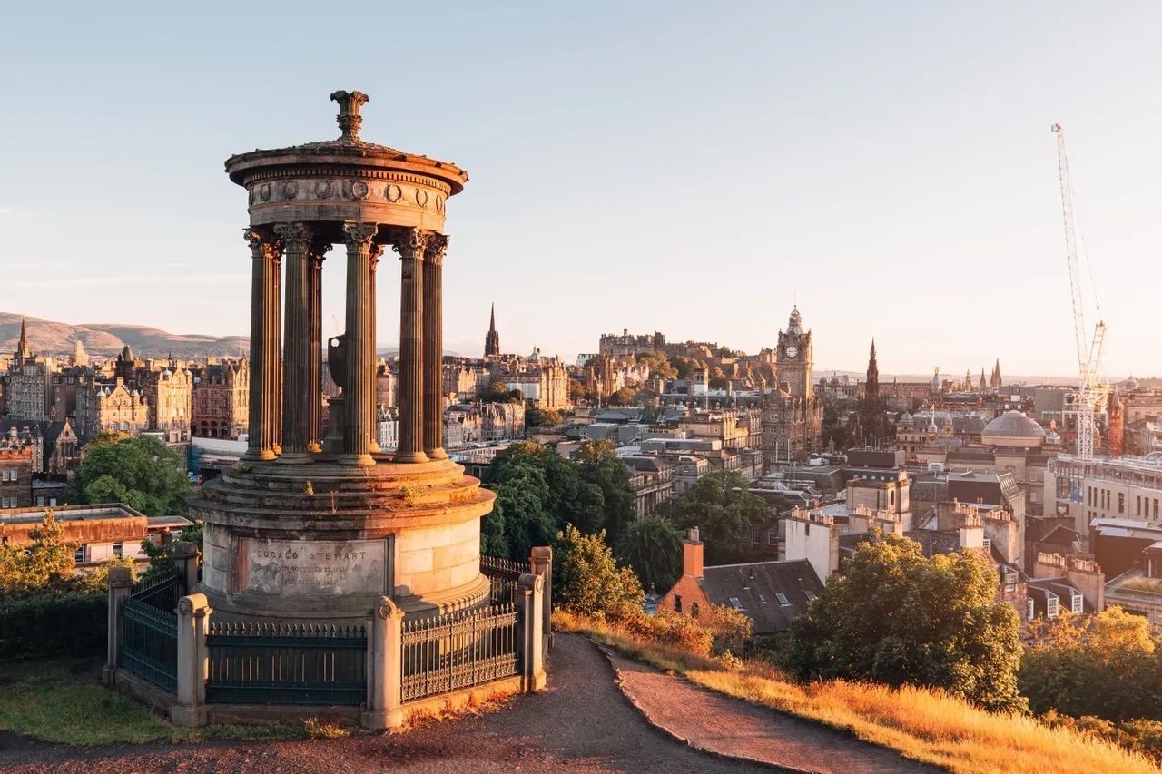 A classical monument on a hill overlooking a historic cityscape with a clock tower during a golden sunset.