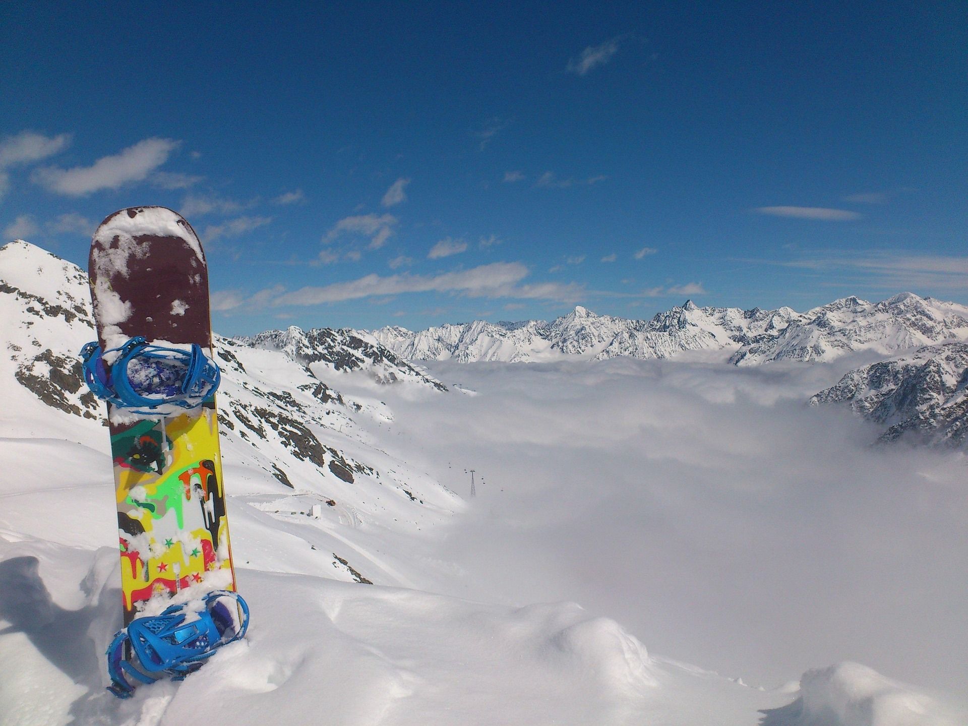 A colorful snowboard stands upright in the snow on a mountain, overlooking a sea of clouds and snow-capped peaks under a blue sky.