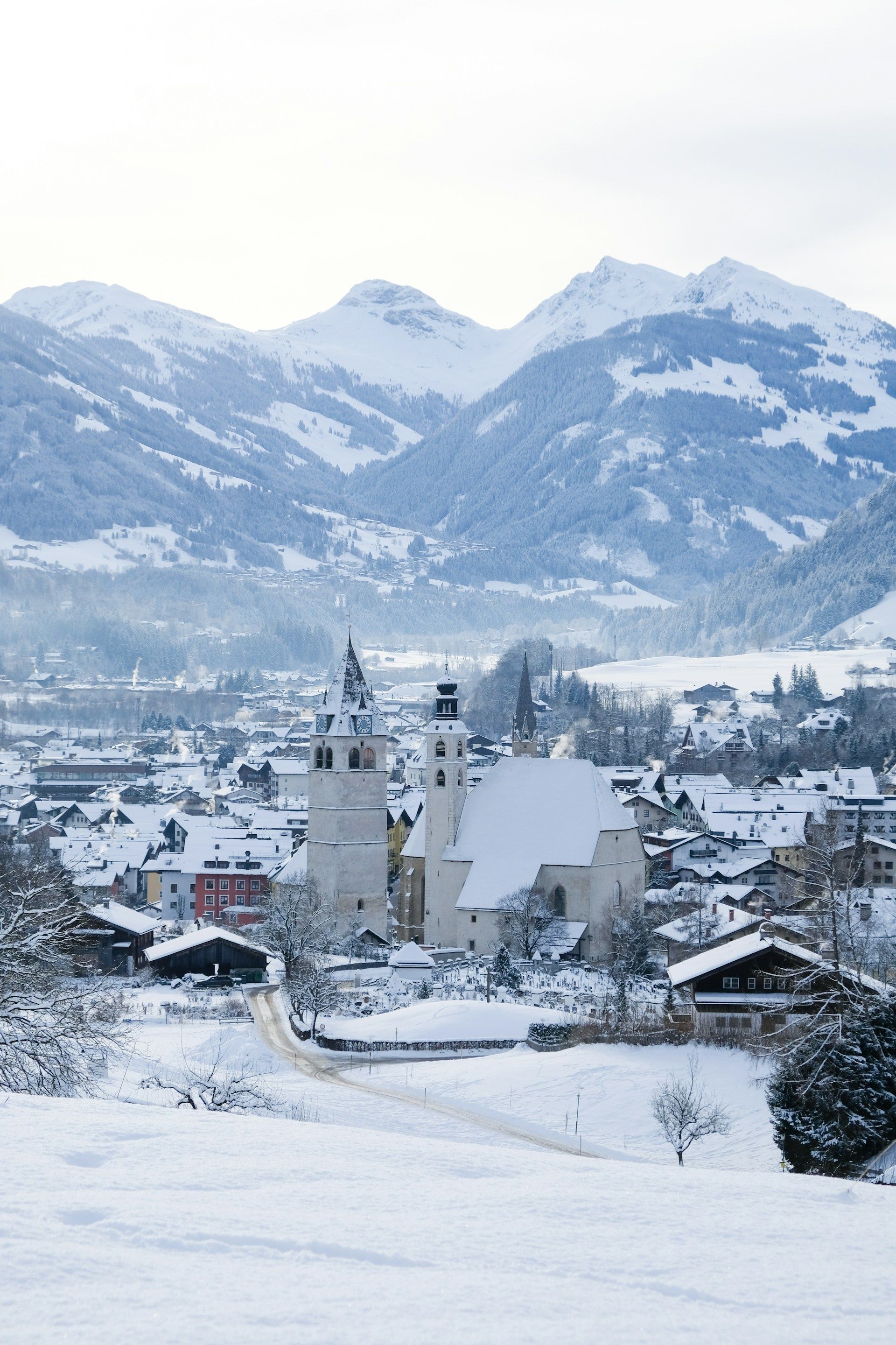 A high-angle view of a snow-covered alpine village with a prominent church, set against a backdrop of snowy mountains.