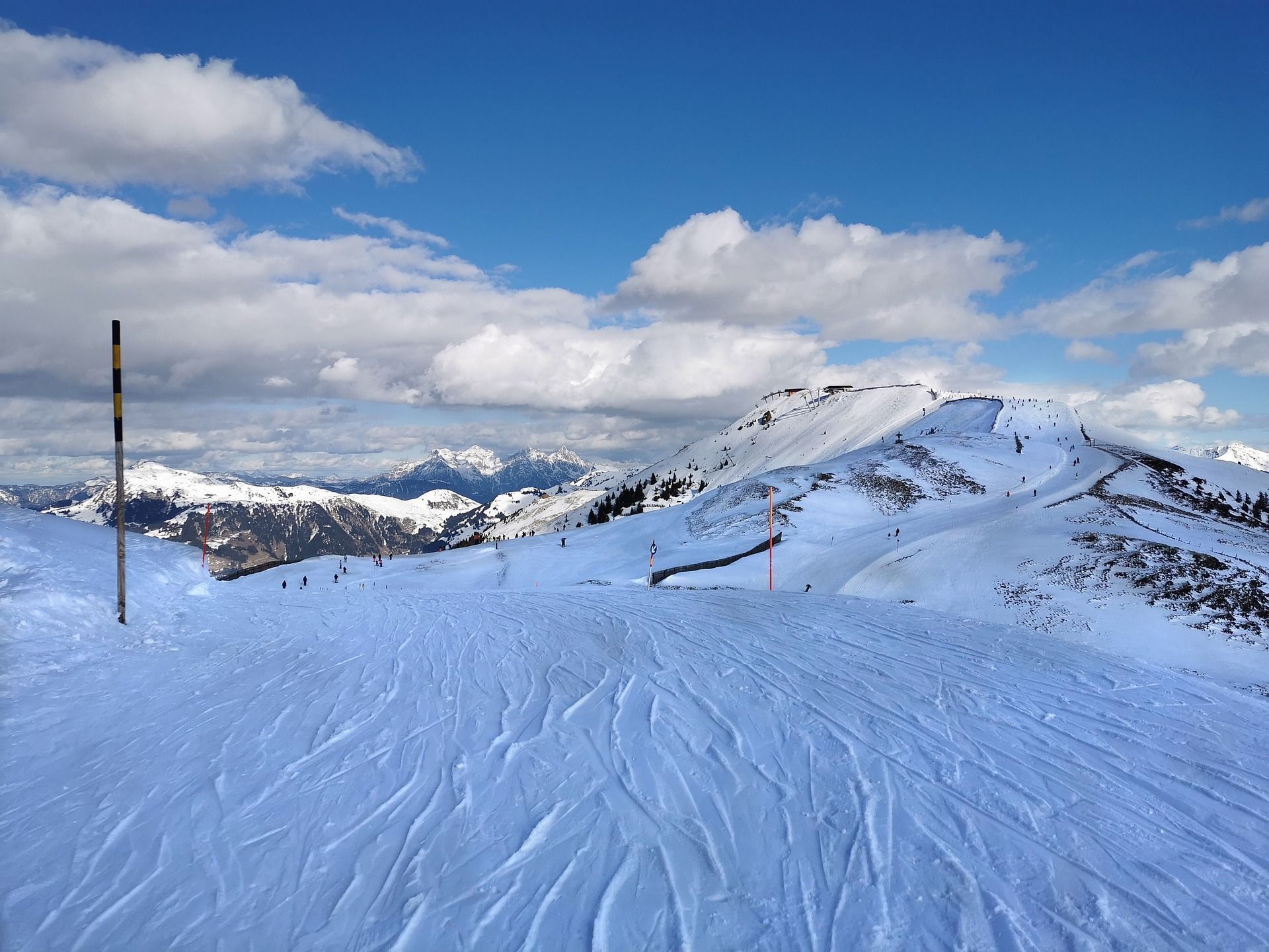 Ein schneebedeckter Berghang mit Skispuren im Vordergrund und entfernten Skifahrern unter blauem Himmel mit weißen Wolken.