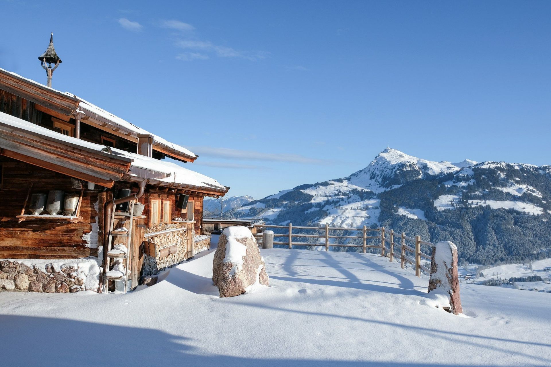 A wooden mountain cabin with a stone foundation is covered in snow, overlooking a snow-capped mountain range under a clear sky.