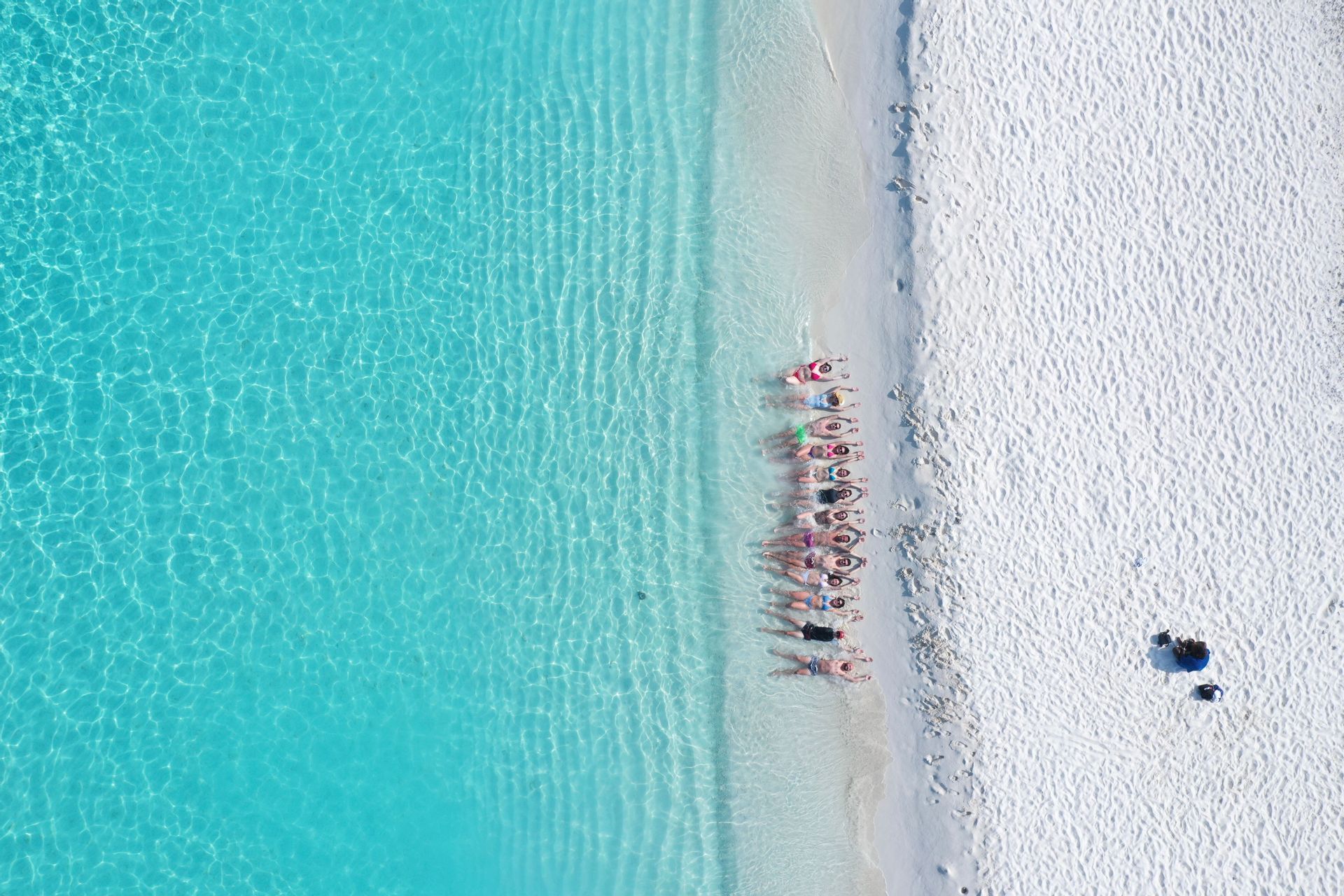 Vista aérea de un grupo de WeRoad tumbado en fila en una playa de arena blanca junto a aguas turquesas y cristalinas.
