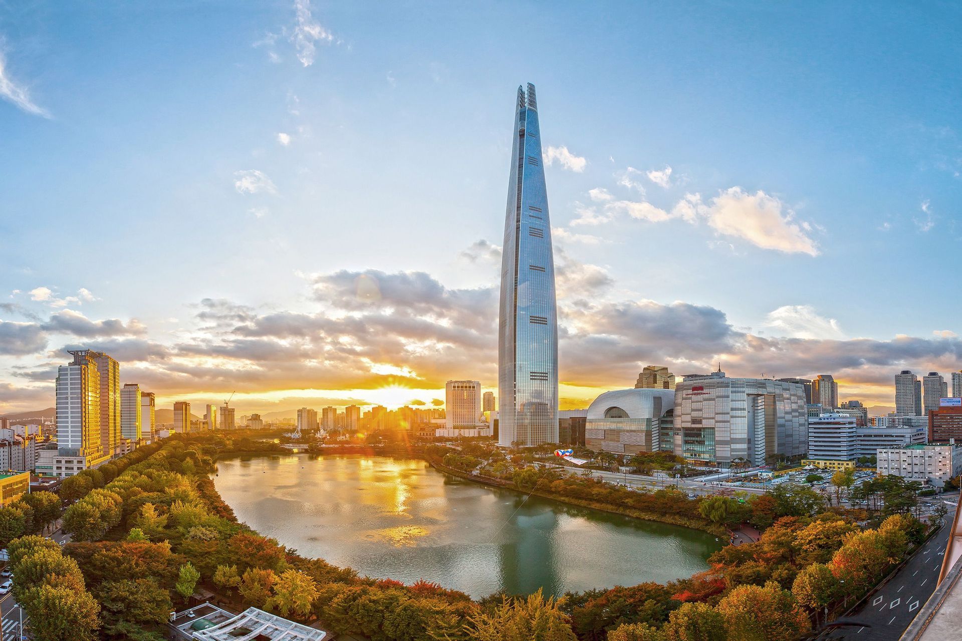 Un alto grattacielo di vetro si erge su uno skyline cittadino accanto a un lago circondato da alberi autunnali al tramonto.