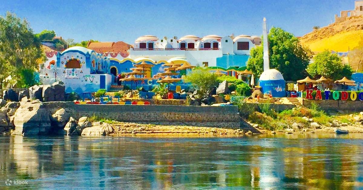 Colorati edifici blu e bianchi in stile nubiano su una sponda rocciosa del fiume, visti dall'acqua.