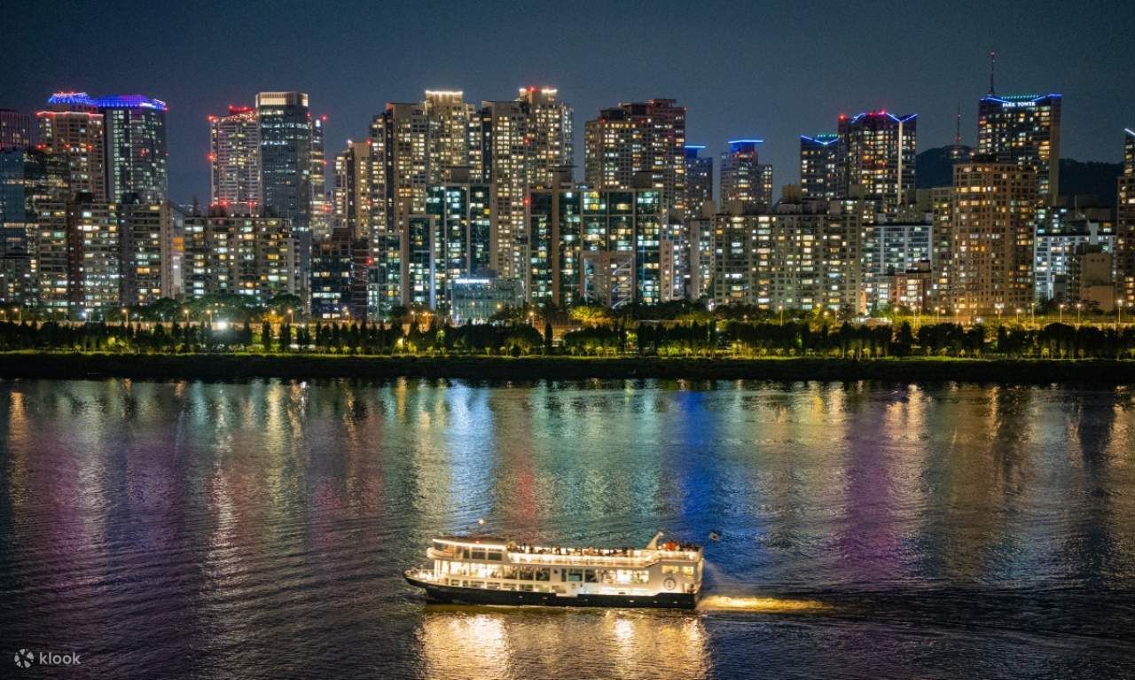 Una barca illuminata naviga di notte su un fiume, di fronte a uno skyline cittadino illuminato.