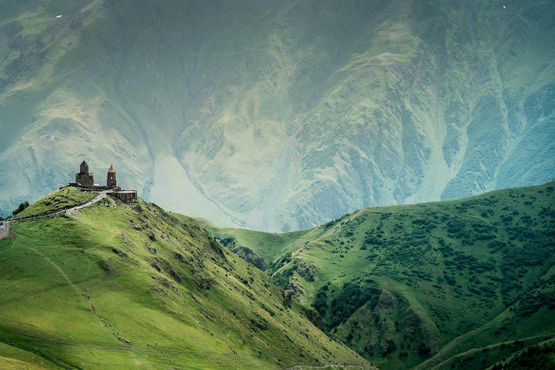 A stone church with two towers sits on a vibrant green hill with massive, hazy mountains in the background.