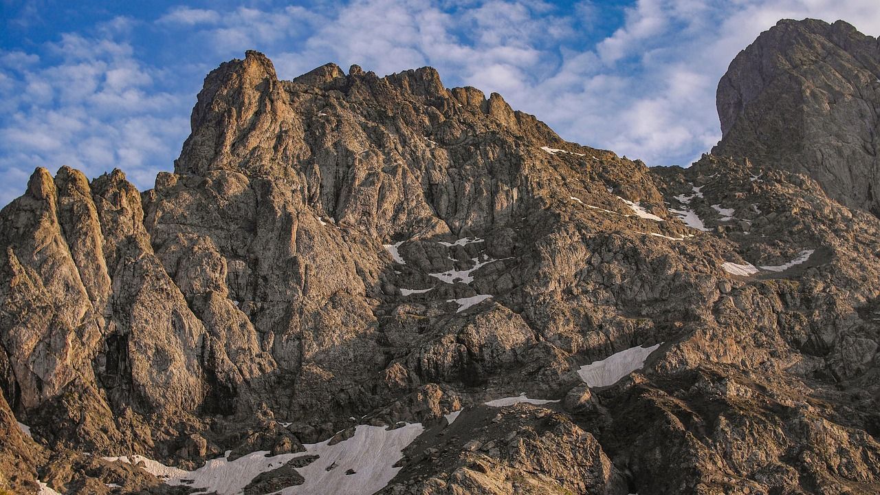 A rugged, rocky mountain peak with patches of snow on its slopes is illuminated by sunlight under a blue, cloudy sky.