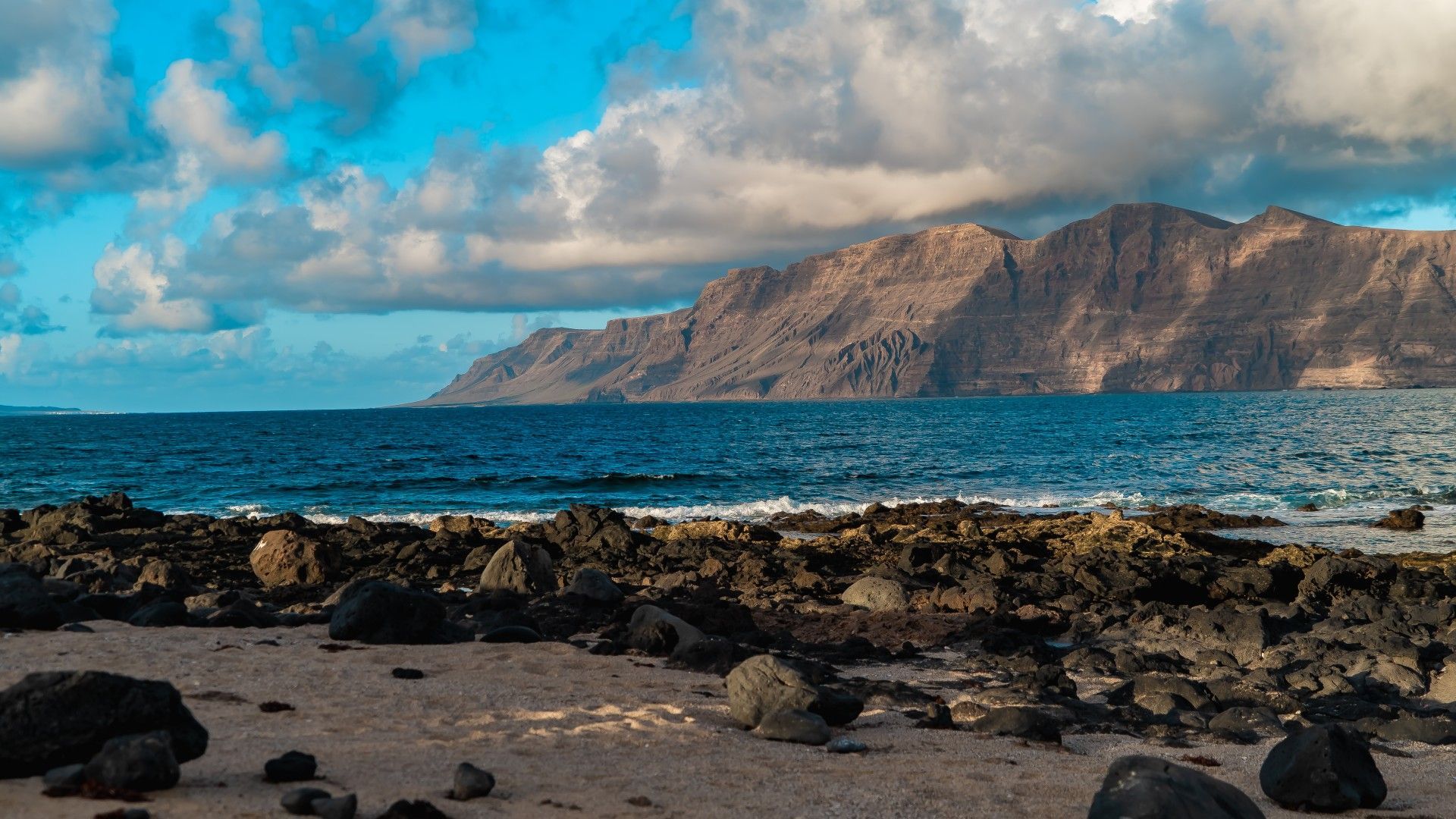 Una costa rocciosa, con sabbia e pietre vulcaniche, si affaccia su una grande catena montuosa oltre l'oceano blu, sotto un cielo parzialmente nuvoloso.