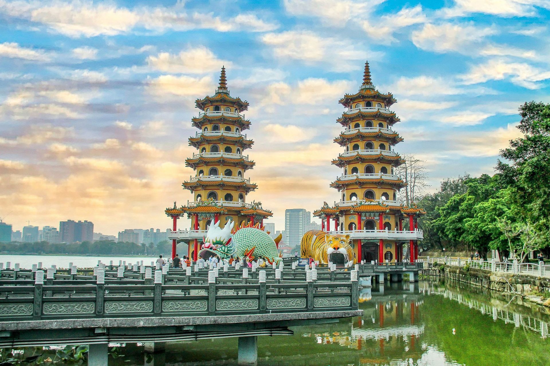 Due pagode ornate con ingressi di drago e tigre si ergono su un lago, con uno skyline cittadino e un cielo nuvoloso sullo sfondo.