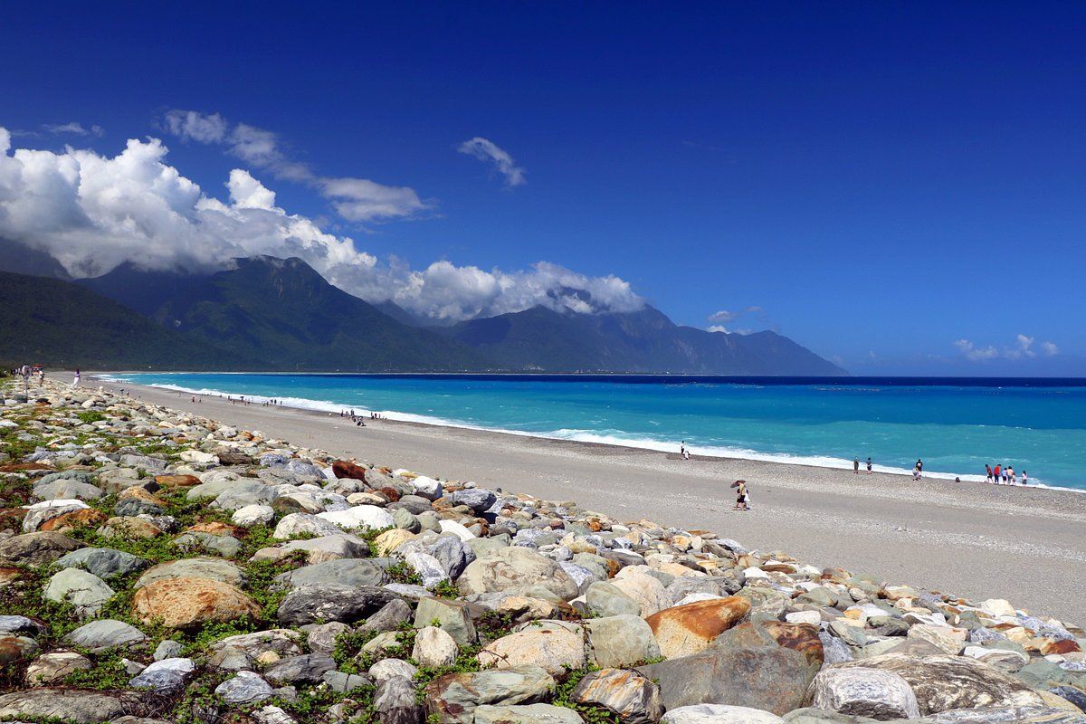 Una costa rocciosa conduce a una spiaggia sabbiosa lungo un mare turchese, con montagne verdi sotto un cielo azzurro parzialmente nuvoloso.