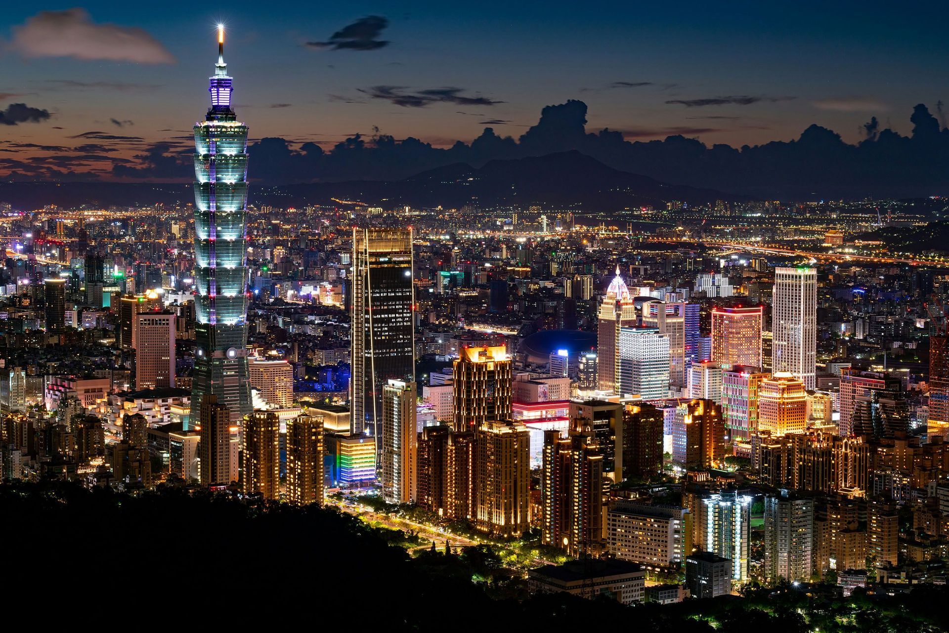 Una vista panoramica di uno skyline cittadino esteso e illuminato di notte, con un alto e distintivo grattacielo che si erge sopra gli altri edifici.