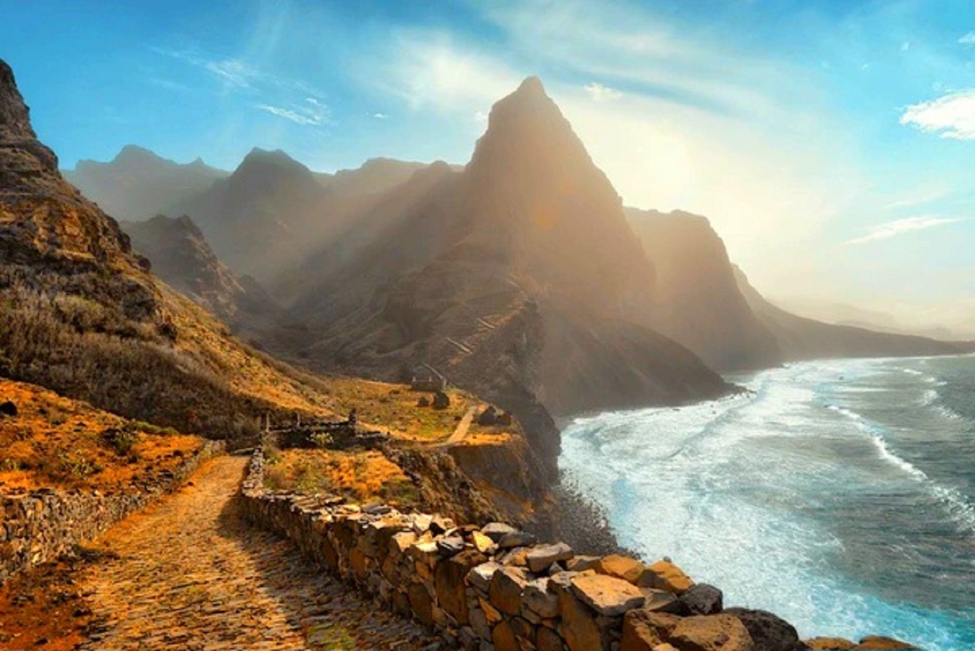Ein Kopfsteinpflasterweg schlängelt sich entlang einer Klippe mit Meerblick, dahinter erheben sich schroffe Berge unter sonnigem Himmel.
