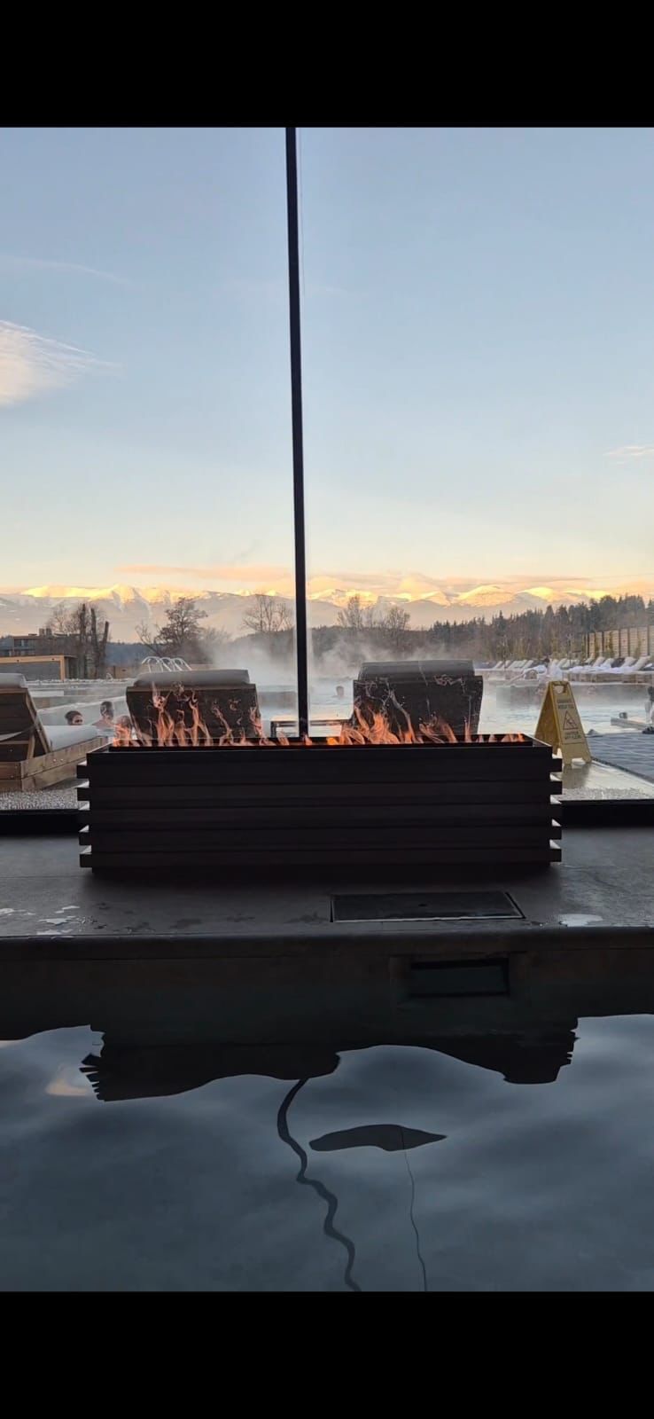 A fire pit burns beside a steaming outdoor thermal pool, with distant snow-capped mountains visible at dusk.