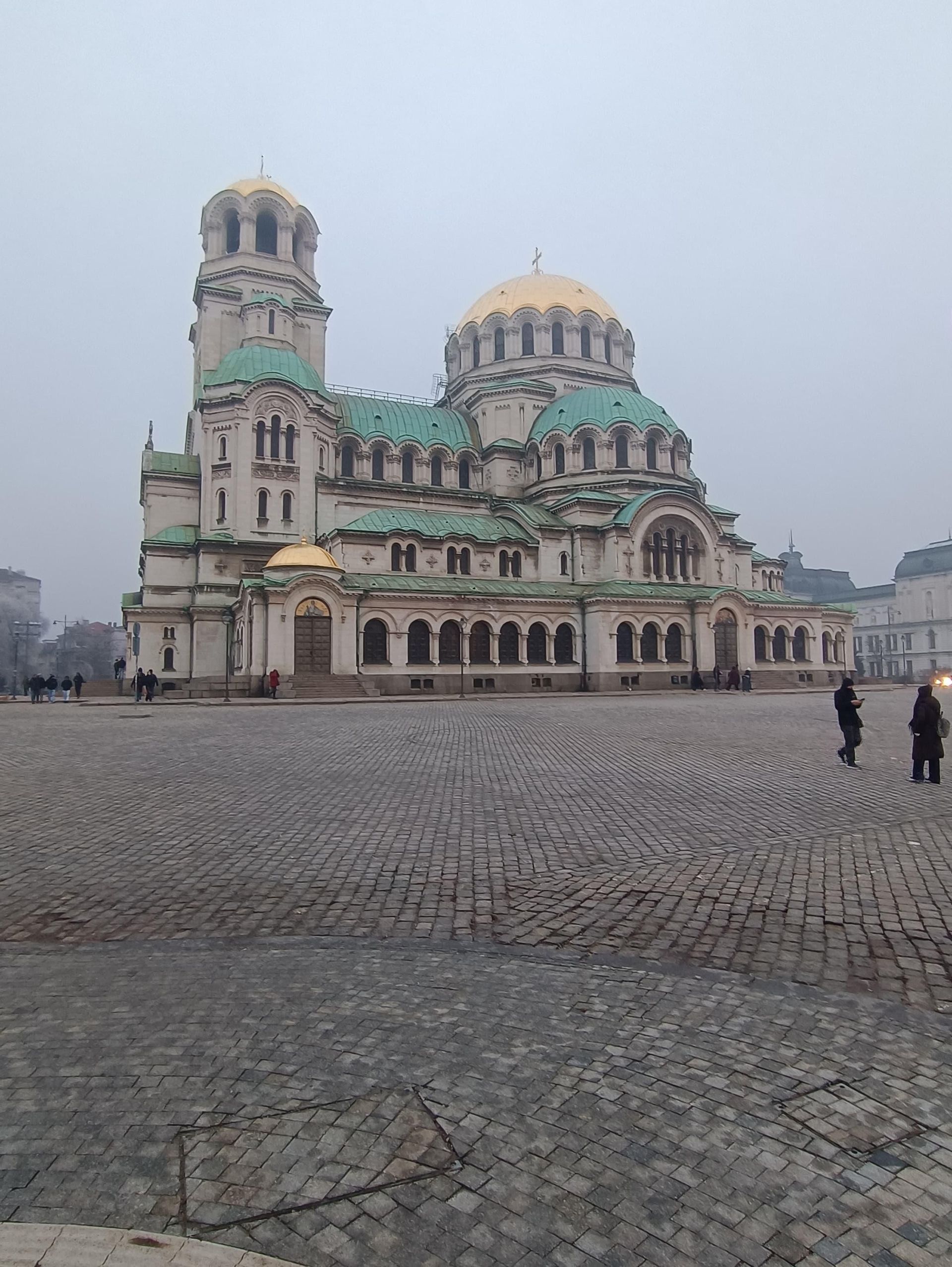 Une cathédrale ornée avec des dômes dorés et des toits verts se dresse sur une grande place pavée par temps de brouillard.