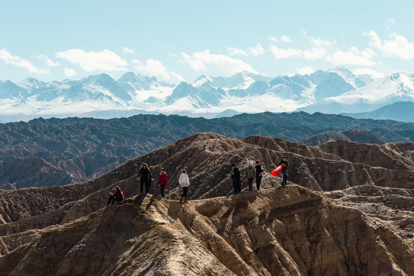 A WeRoad group trip stands on a ridge overlooking a vast canyon landscape, with a distant snow-capped mountain range under a blue sky.