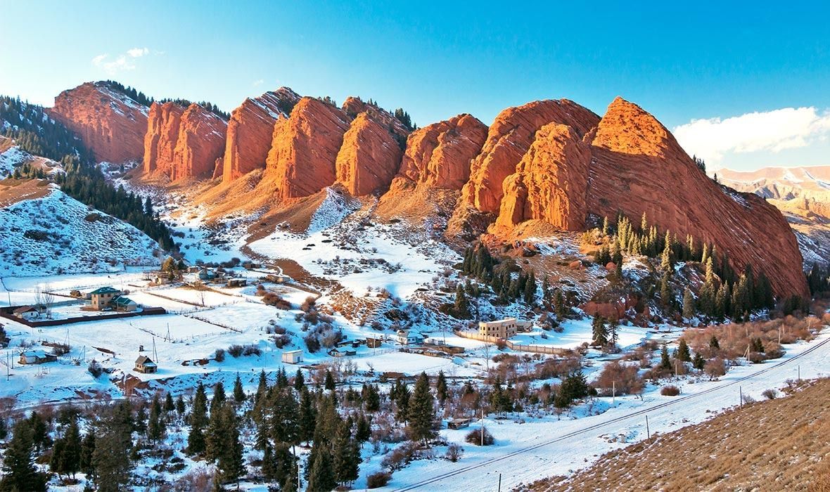 Sunlight illuminates large red rock formations towering above a snow-covered valley dotted with pine trees and a small village.
