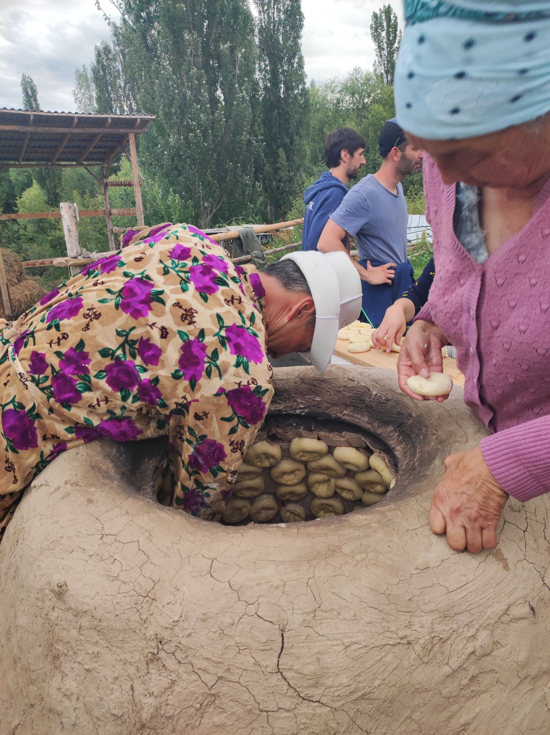 Un homme se penche vers un grand four tandoor en argile pour étaler de la pâte à pain sur ses parois, observé par une femme et un groupe WeRoad en déplacement.