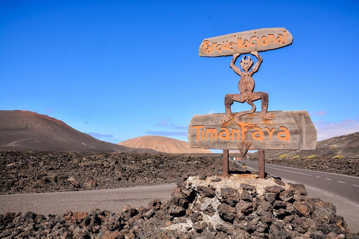 Un cartello di legno per il Parco Nazionale di Timanfaya con il suo simbolo "El Diablo" si trova su rocce vulcaniche accanto a una strada in un paesaggio di campo lavico.