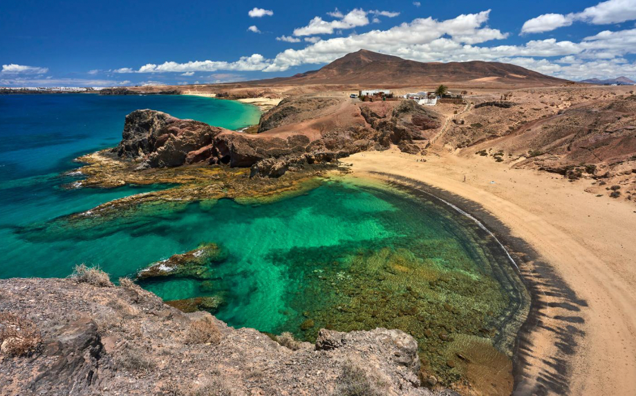 Vista dall'alto di una caletta appartata con acque cristalline turchesi, una spiaggia di sabbia e un paesaggio roccioso sotto un cielo azzurro.
