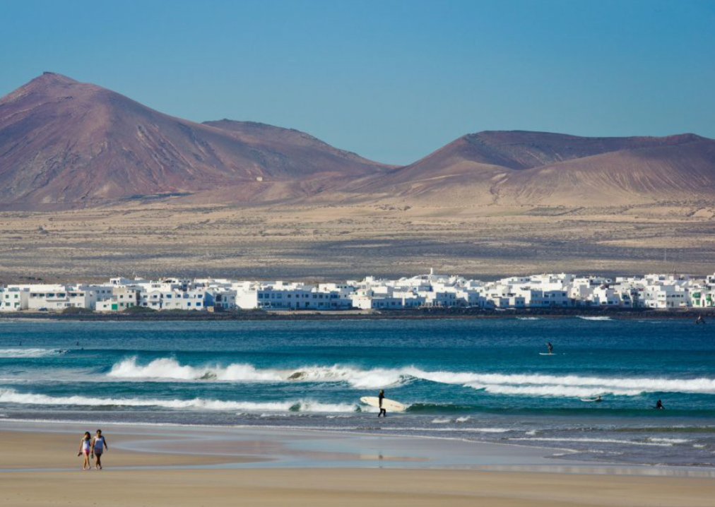 Una spiaggia sabbiosa con persone e surfisti nell'oceano, con una città costiera bianca e montagne aride sullo sfondo.