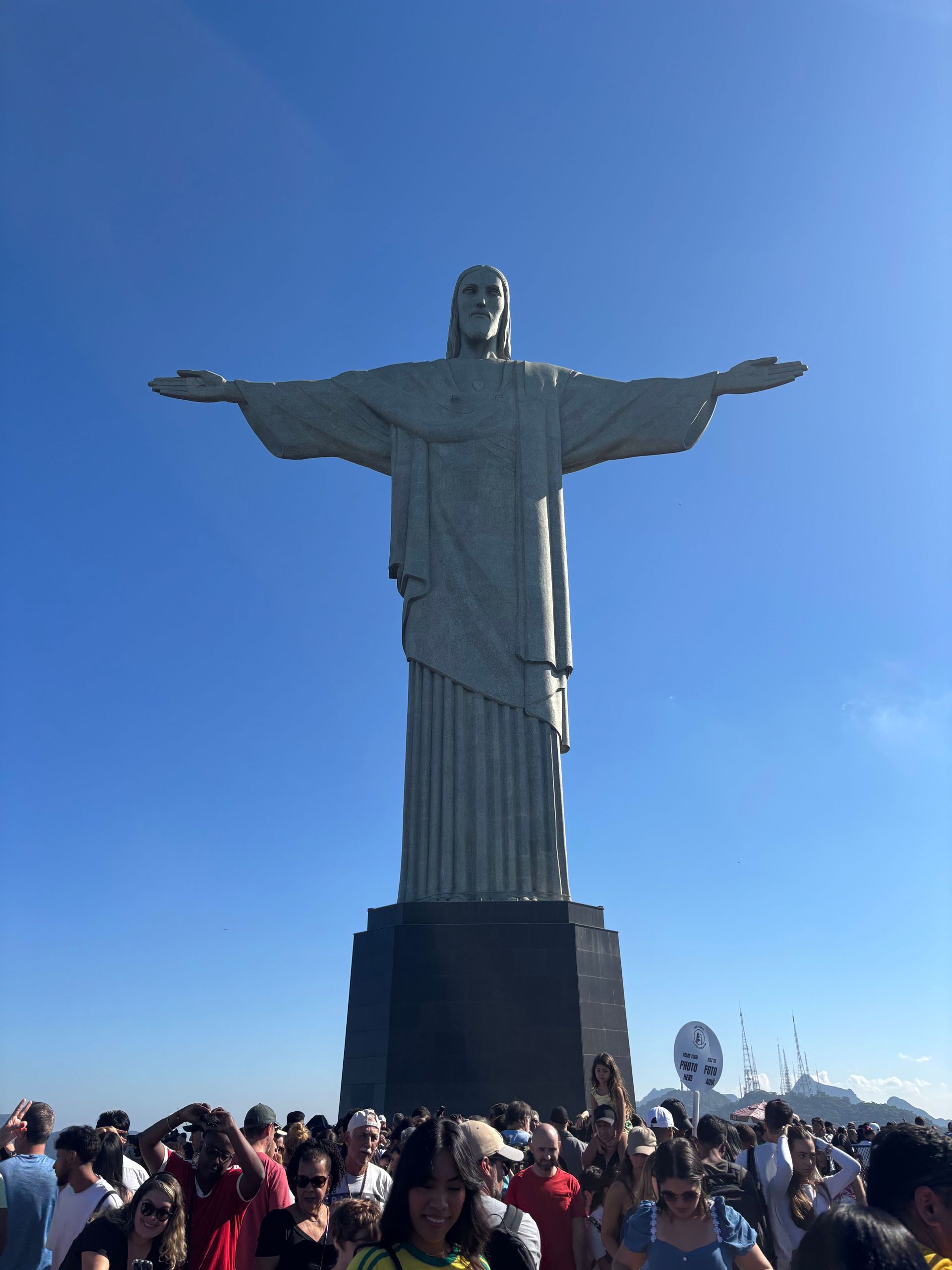 La statua del Cristo Redentore si erge imponente su una folla di persone alla sua base, contro un cielo azzurro e limpido.