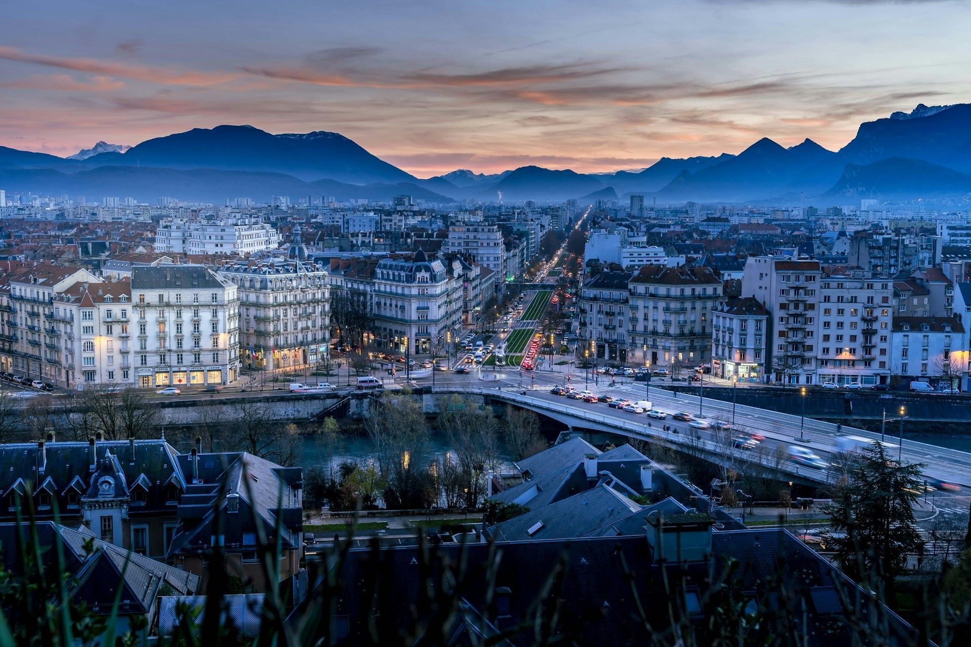 Vue aérienne d'une ville au coucher du soleil, avec des rues et des bâtiments illuminés et une chaîne de montagnes en arrière-plan.