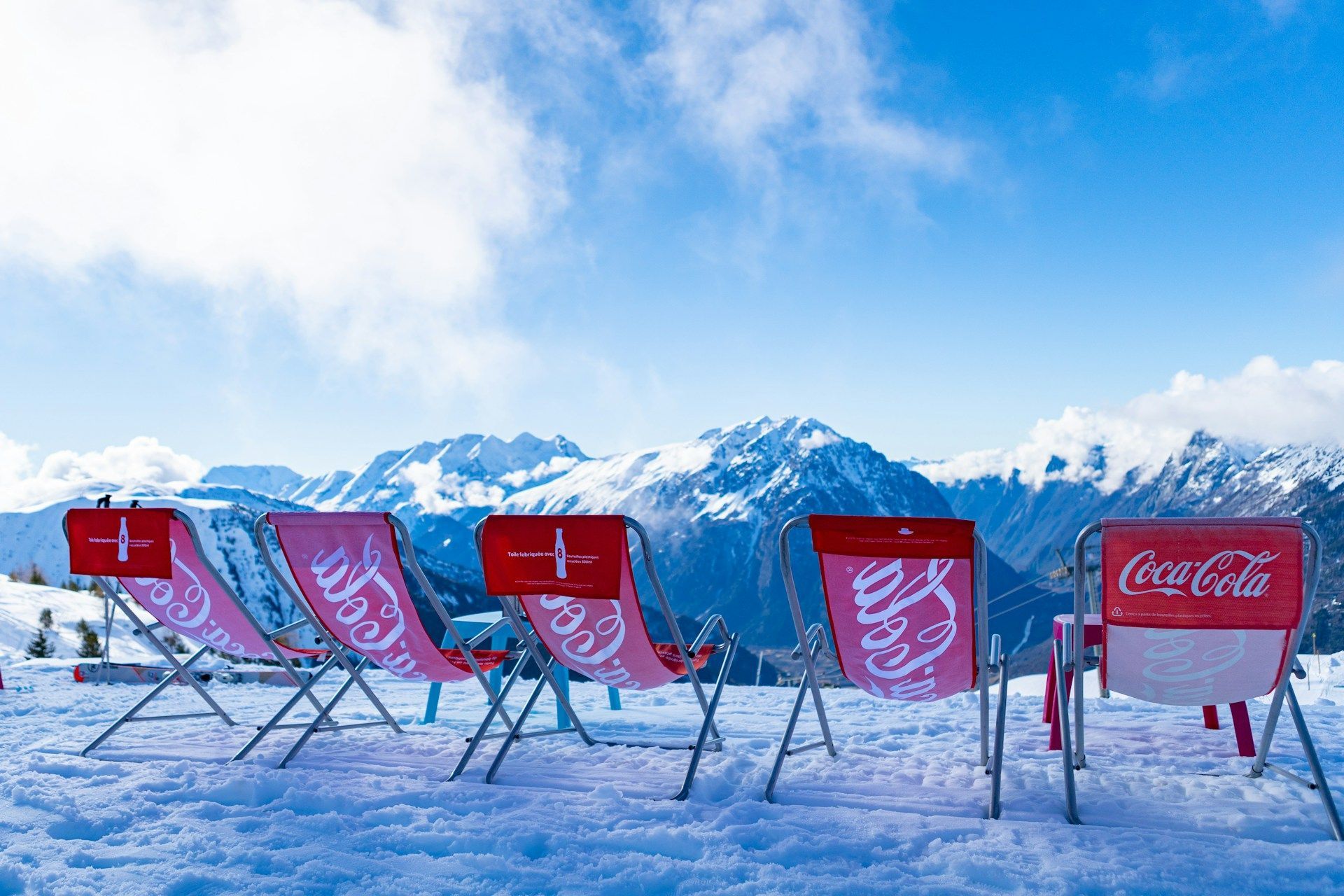 A row of red Coca-Cola branded deck chairs sit empty in the snow, facing a mountain range under a blue sky.
