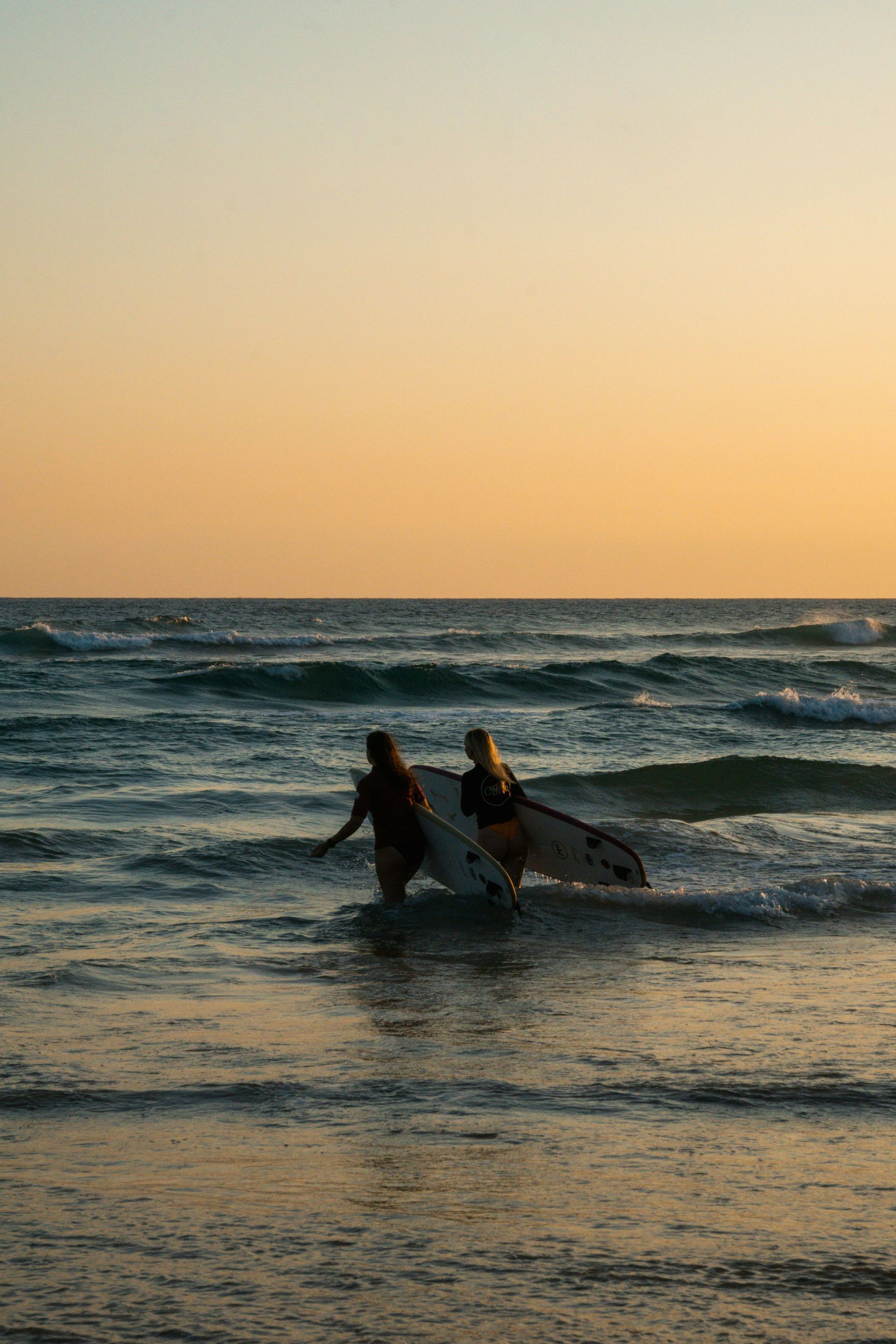 Due donne con tavole da surf camminano tra le onde dell'oceano sotto un caldo cielo al tramonto.