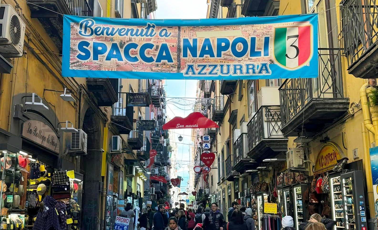 Un grande striscione blu per 'Spaccanapoli Azzurra' pende su una strada stretta e affollata fiancheggiata da negozi ed edifici gialli.