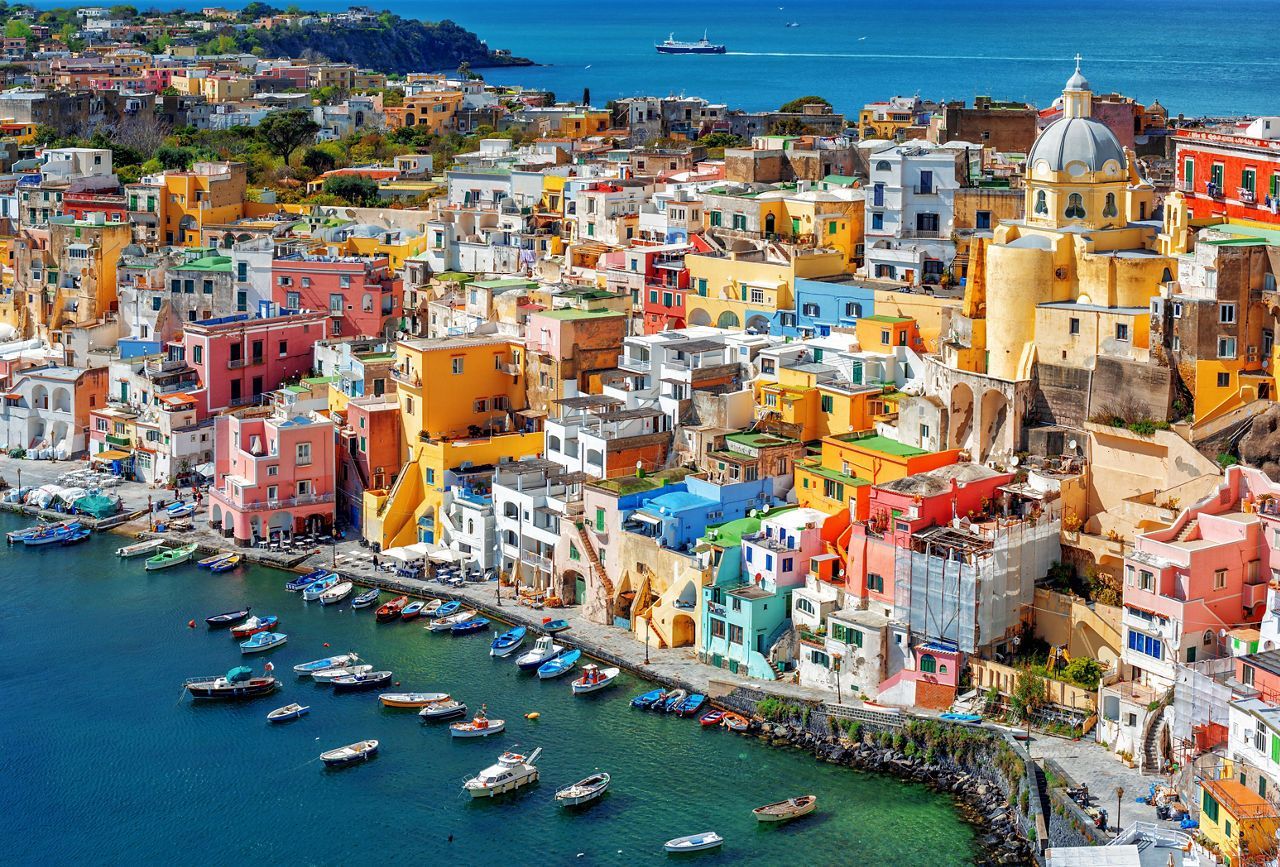 Edifici colorati di una città di mare sono fitti su una collina che domina un porto pieno di piccole barche.
