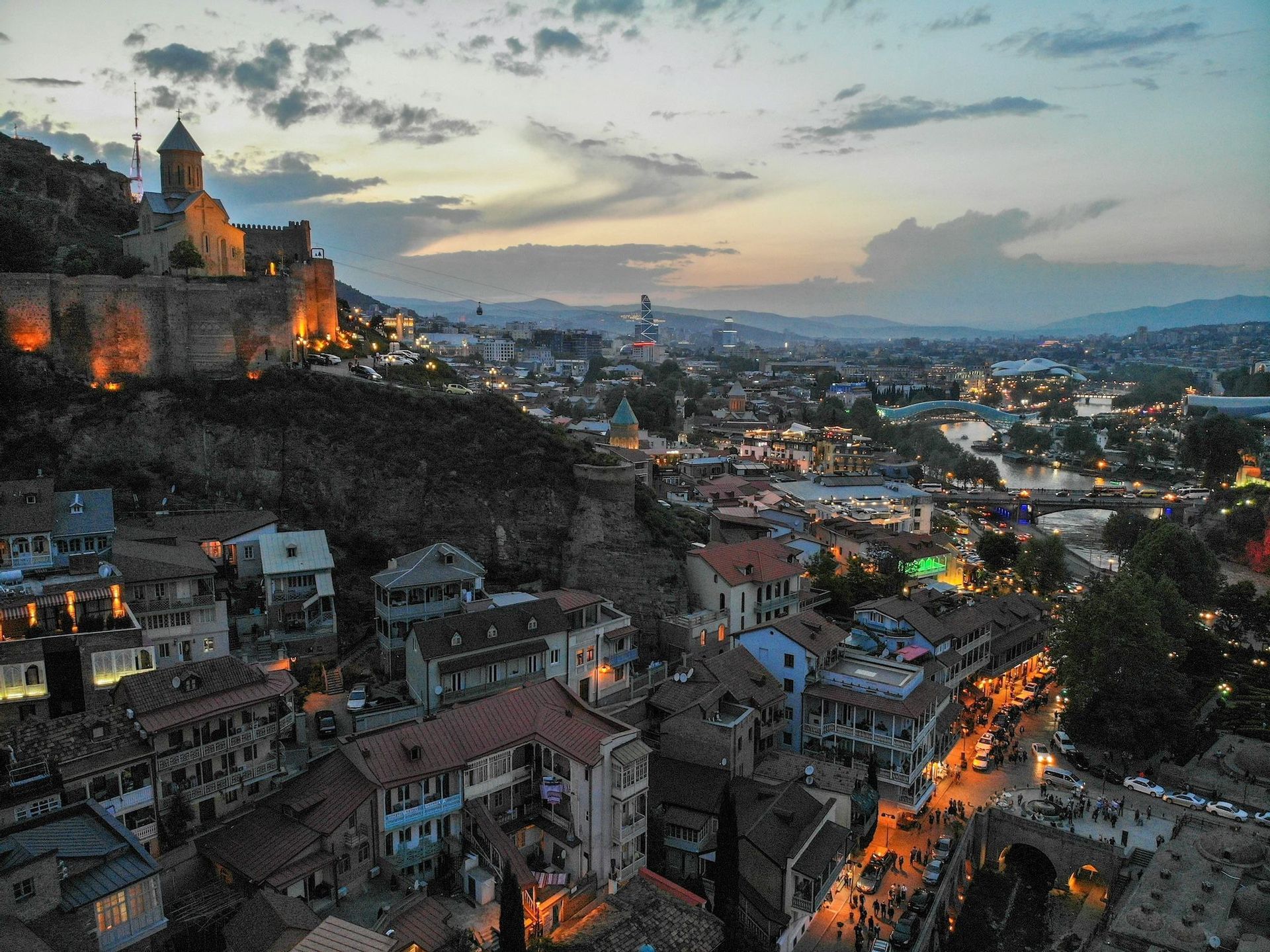 An ancient city with an illuminated fortress on a hill overlooking a river and lively streets at sunset.